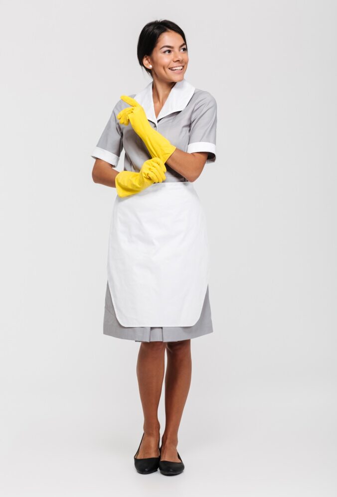 Full length portrait of a attractive smiling housekeeper dressed in uniform posing while standing and looking away isolated over white background