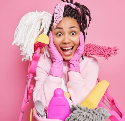 Impressed surprised cheerful Afro American woman grabs face supplies with cleaning service surrounded by equipment necessary for tidying up room isolated over pink background. Domestic work.